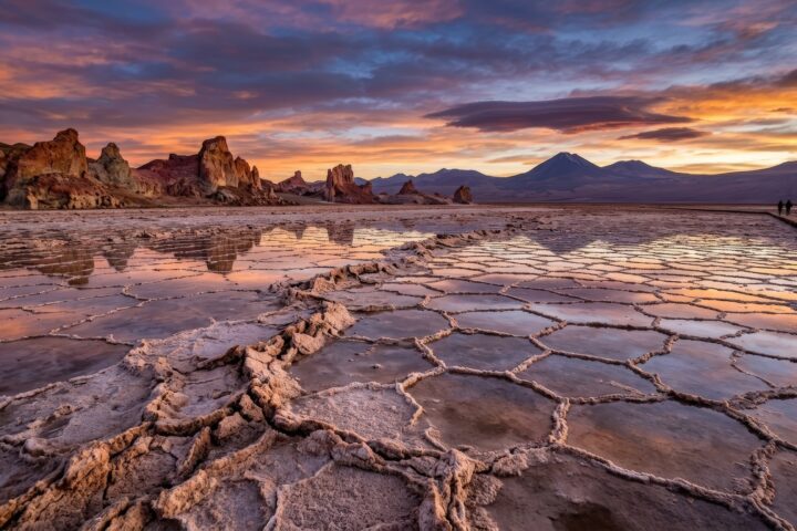 Salt Flats With Hexagonal Crust Formations Reflecting Sunset Sky. Landscape With Woman And Man Walking On Wooden Path In Atacama Desert. Natural Geological Wonder And Travel Scenery.
