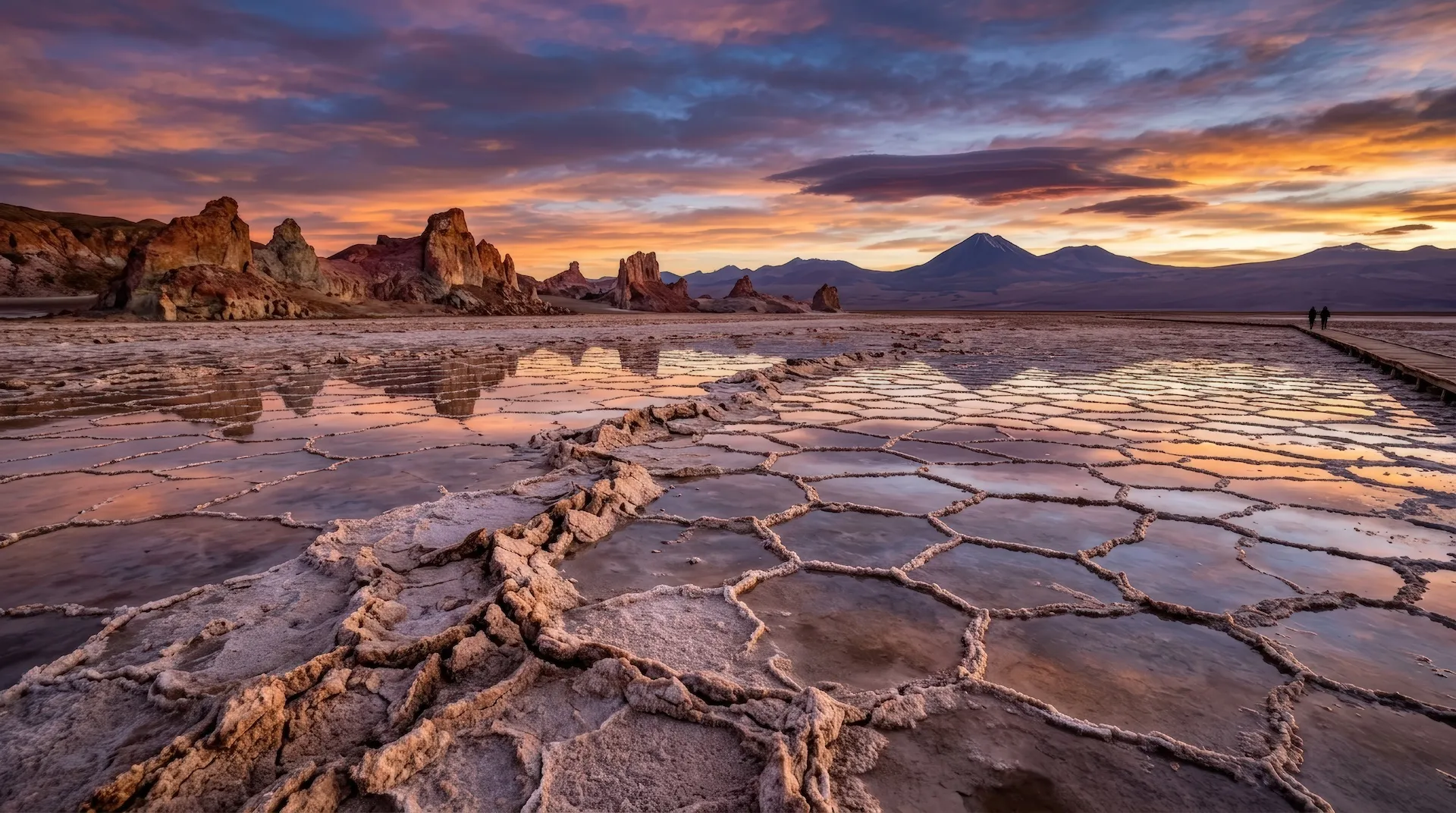Salt Flats With Hexagonal Crust Formations Reflecting Sunset Sky. Landscape With Woman And Man Walking On Wooden Path In Atacama Desert. Natural Geological Wonder And Travel Scenery.